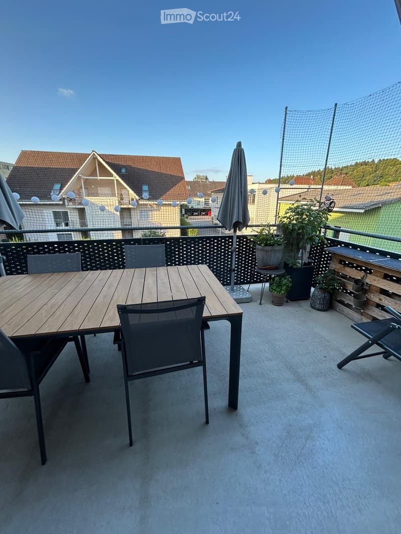 Balcony with a wooden table, black chairs, umbrella, and potted plants, overlooking a residential area.