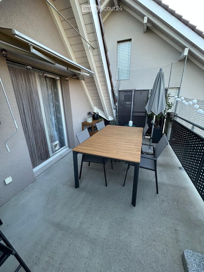 Balcony with a wooden table, chairs, and an umbrella, featuring a small shelf with a teapot and cup, and a view of the building exterior.