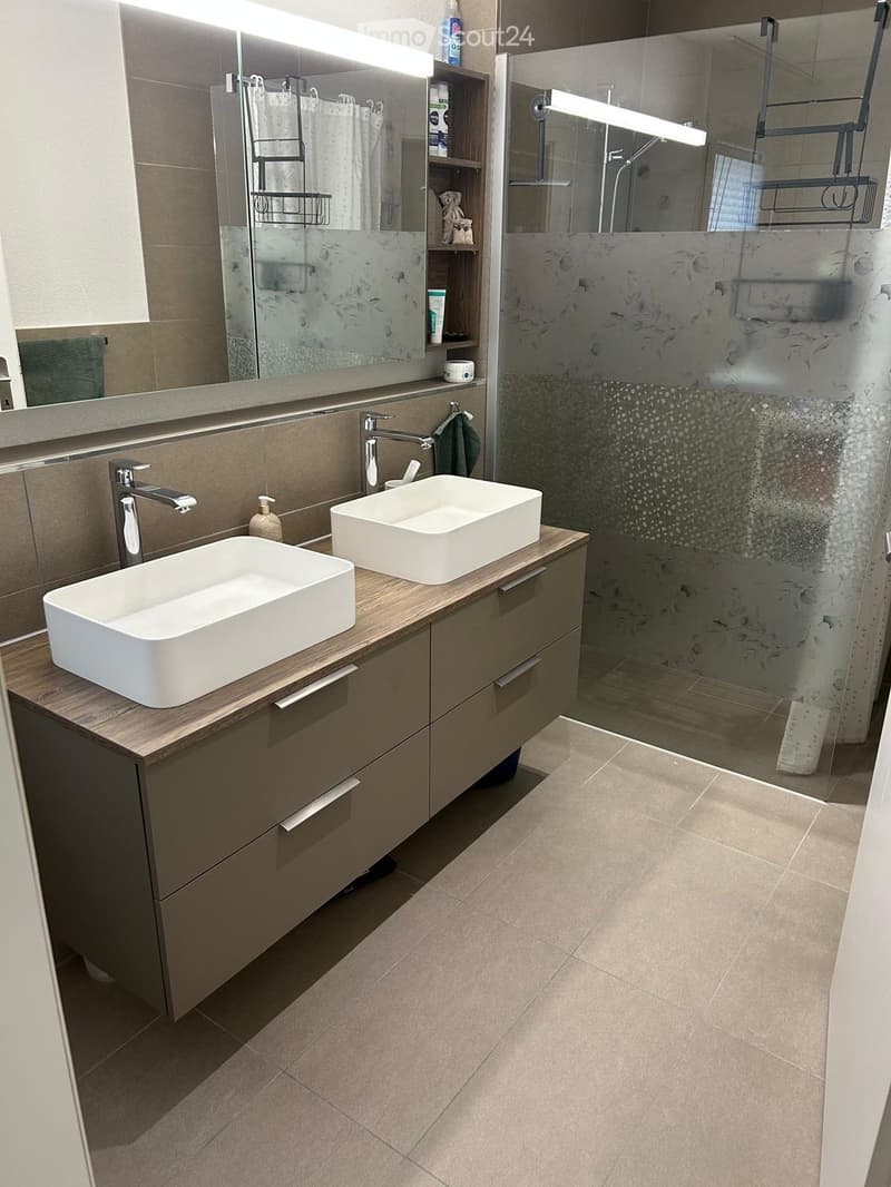 Modern bathroom with two white sinks on a wooden vanity, a large mirror with built-in lighting, and a glass-enclosed shower area.