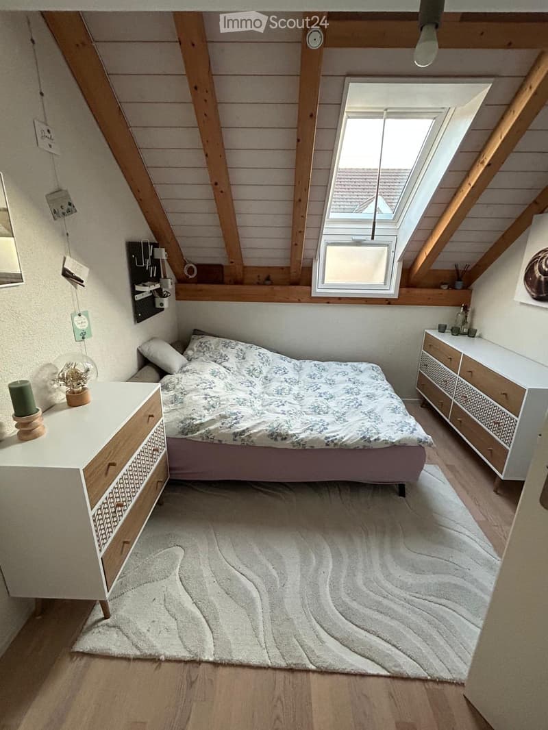 Cozy attic bedroom with wooden beams, slanted ceiling, skylight window, bed with patterned bedding, nightstands, and decorative items.