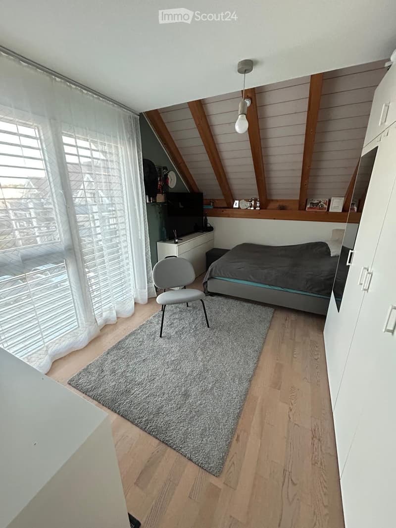Modern attic bedroom with wooden slanted ceilings, large window with white curtains, grey bed, grey chair, grey rug, white cabinet, and TV on a shelf.