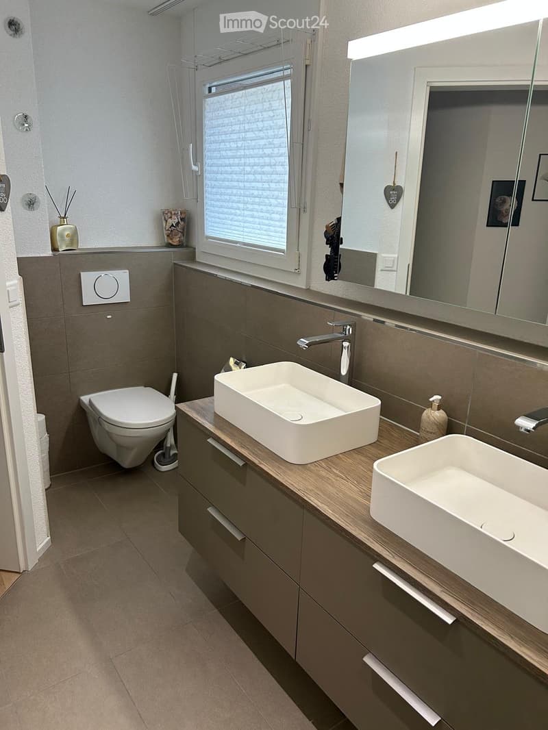 Modern bathroom with a toilet, two sinks, a large mirror, and a window with blinds. The cabinetry is grey with wooden accents, and the floor is tiled.