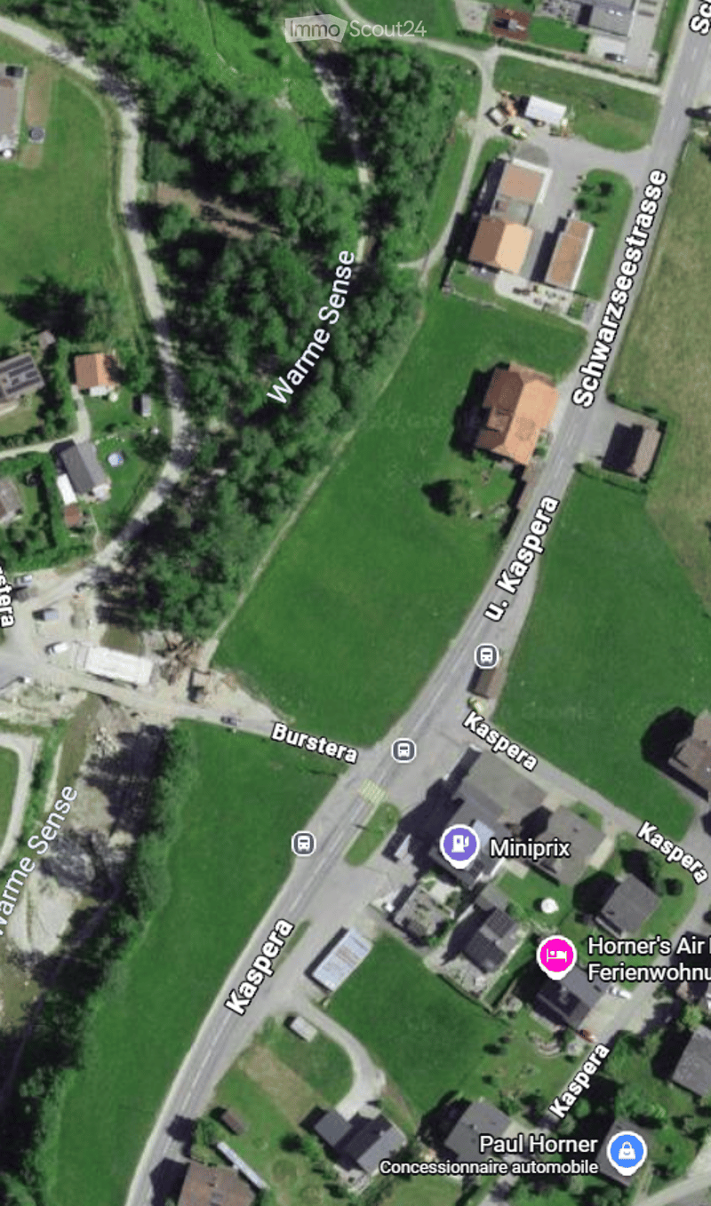 Aerial view of a rural area with green fields, roads, and scattered buildings including houses and a parking lot.