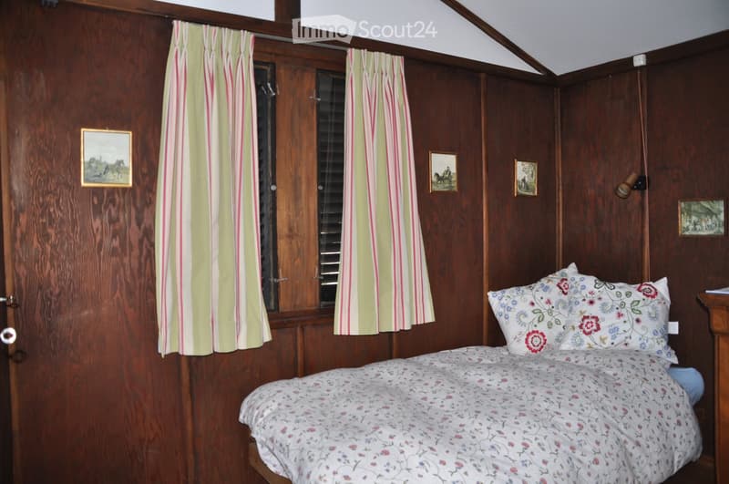 Cozy bedroom with wooden walls, a bed with floral bedding, striped curtains, and several framed pictures on the walls.