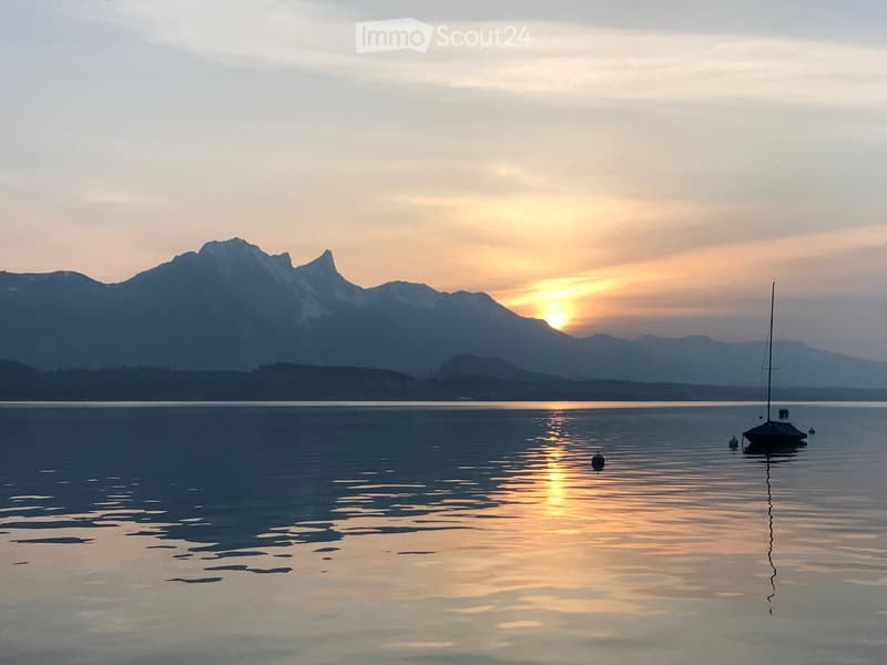 Serene lakeside scene at sunset with calm water reflecting the golden light, distant mountains silhouetted against the sky, and a small sailboat anchored near the shore.
