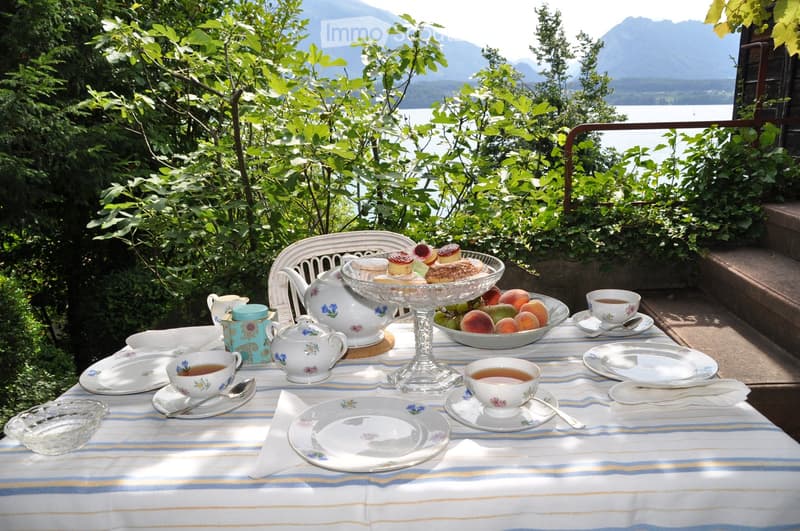A beautifully set tea table on a balcony overlooking a lake and mountains, with teapots, cups, plates, cakes, and a bowl of fruit.