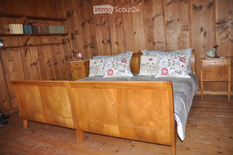 Cozy bedroom with wooden walls and floor, featuring a wooden bed with floral pillows and a nightstand with a table lamp. Shelves with books are mounted on the wall.