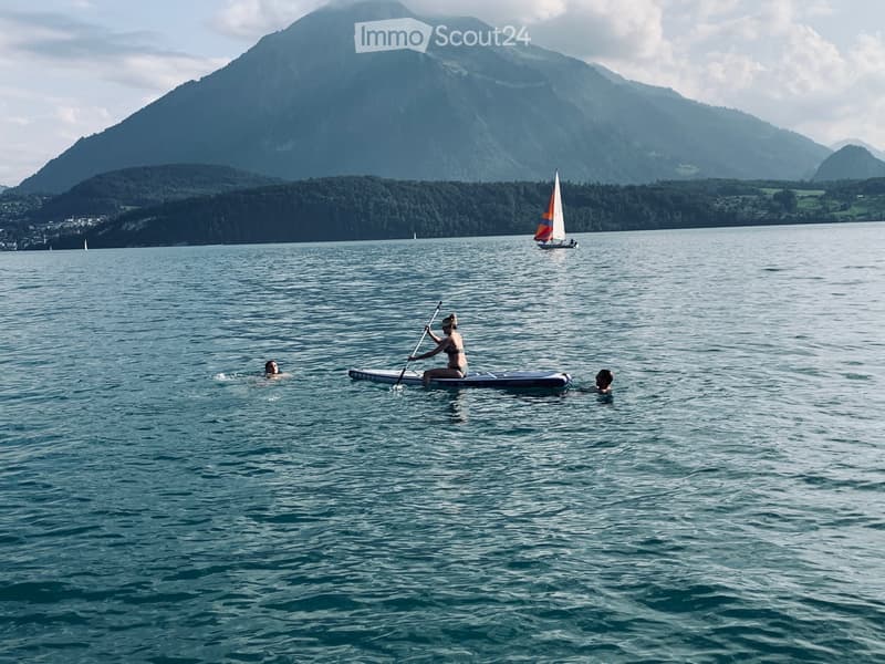 A serene lake scene with a person paddleboarding and two individuals swimming, with a sailboat in the background and a mountain range visible.