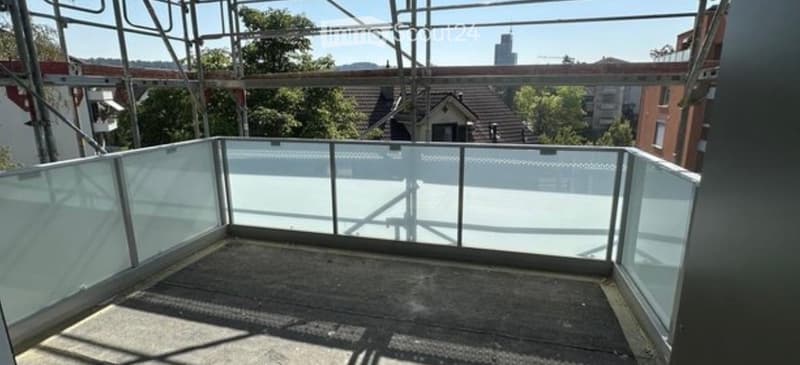 Empty balcony with glass railings, overlooking a cityscape with buildings and trees.