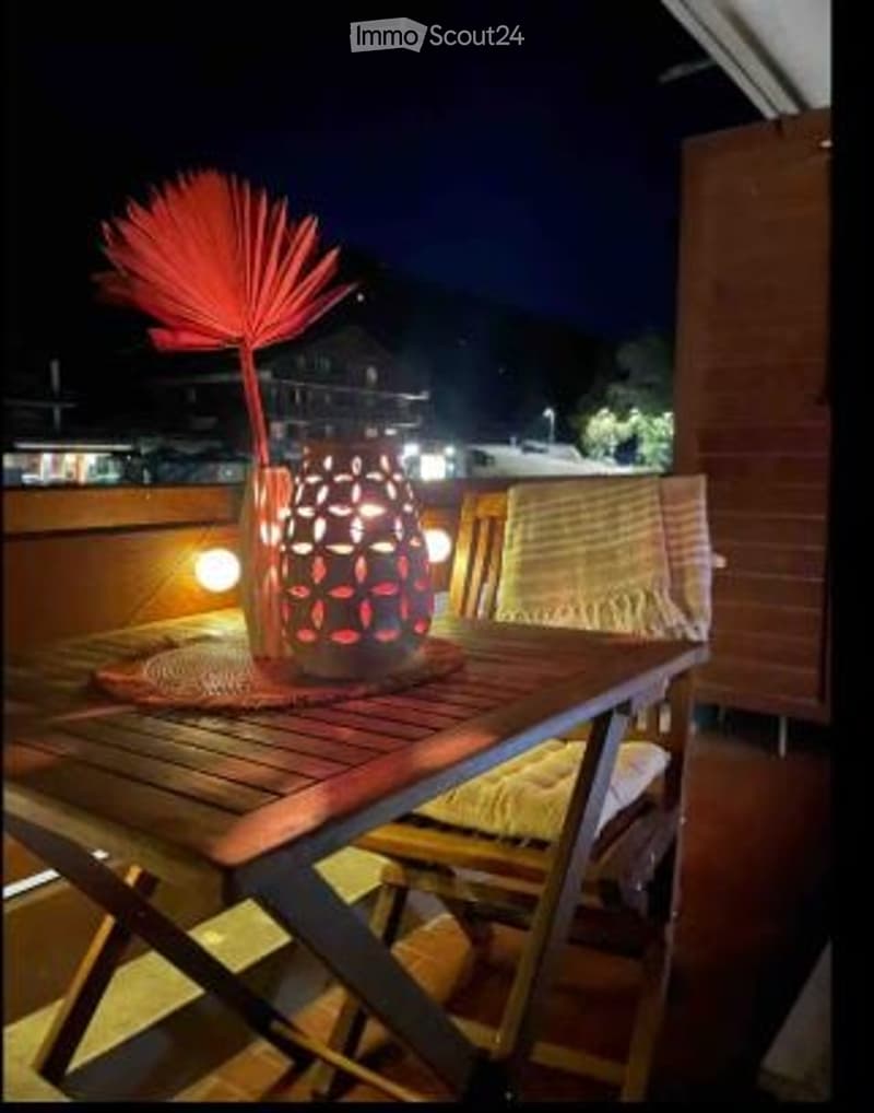 Nighttime balcony with a wooden table, decorative lamp, and a woven chair. The background shows a view of a town with lights.