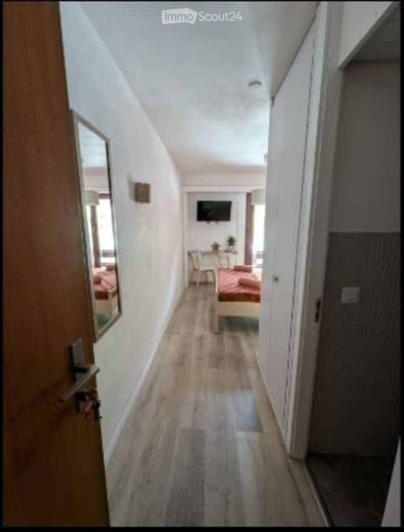 A narrow hallway leads to a small bedroom with a bed, a TV on a stand, a chair, and a small table. The floor is light wood, and the walls are white.