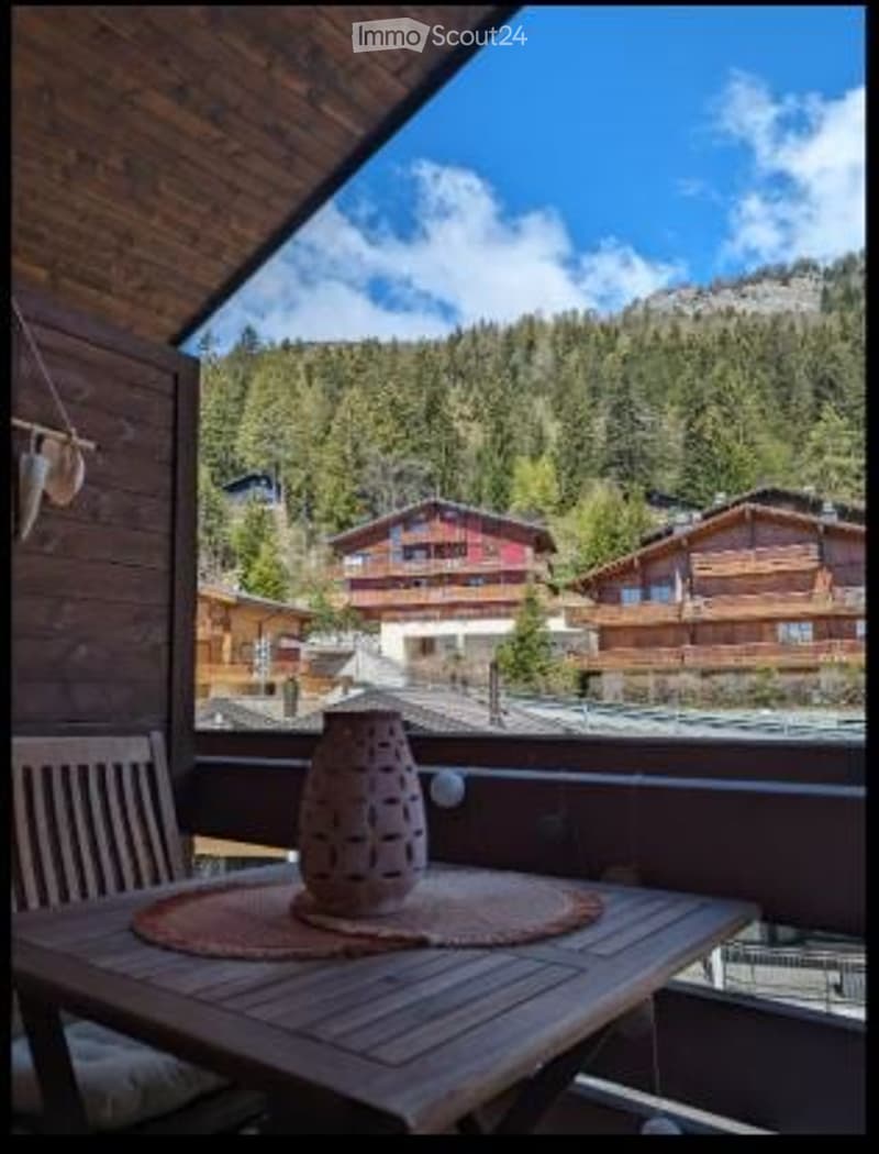 Balcony with wooden table and chair, overlooking a scenic mountain village with wooden houses and lush green trees.