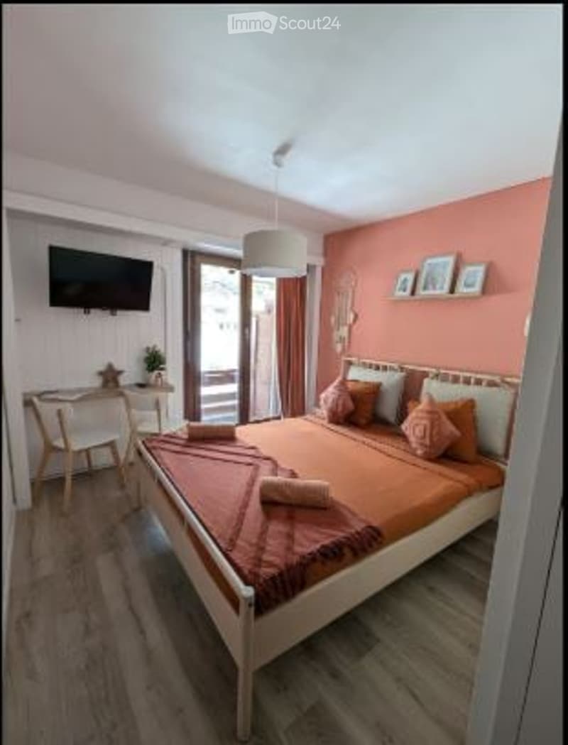 Bright bedroom with a double bed, orange bedding, a nightstand, a chair, a small table, a lamp, a television, and a balcony with a view.