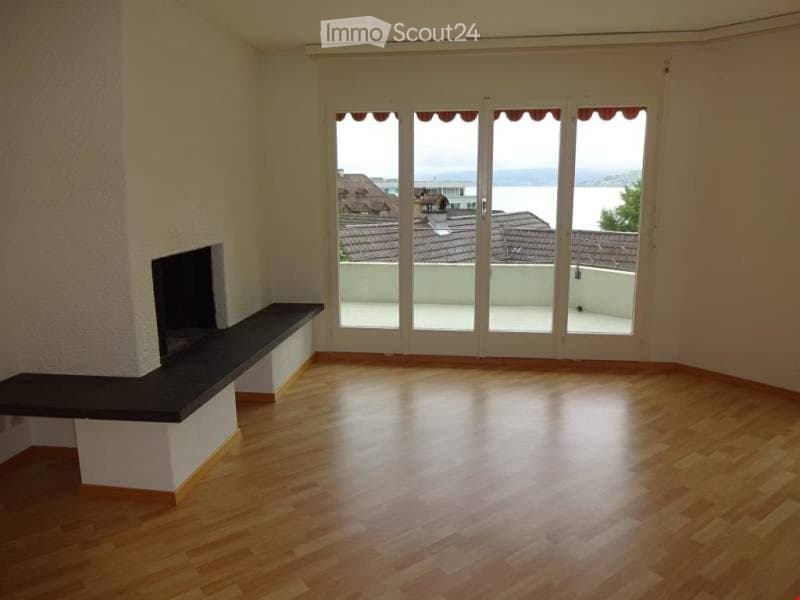 Empty room with wooden flooring, large glass doors leading to a balcony, and a built-in fireplace with a bench.
