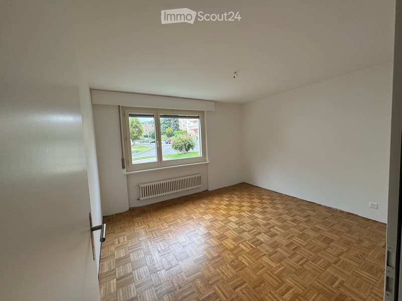 Empty room with wooden herringbone flooring, white walls, a window with a radiator below, and a door visible on the left side.