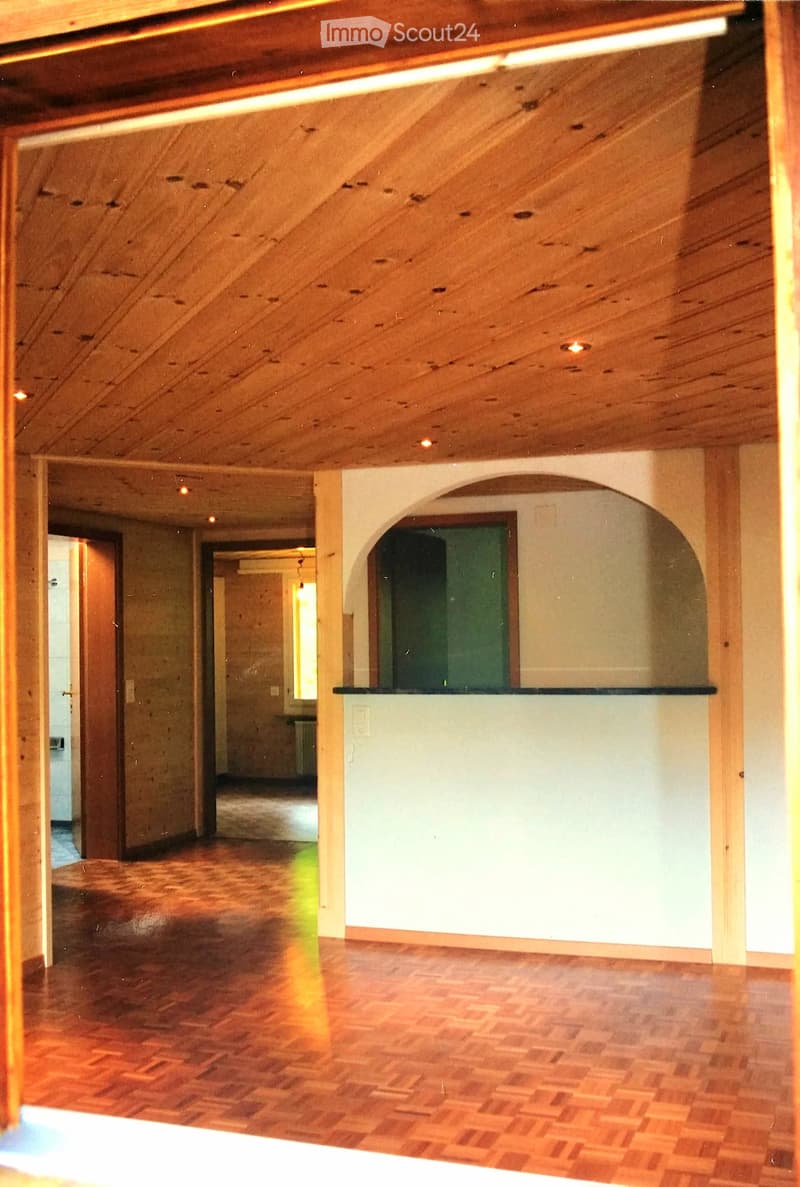 An empty room with wooden floors, exposed wooden beams on the ceiling, and a small counter area with a green door behind it. The space is well-lit with recessed lighting.