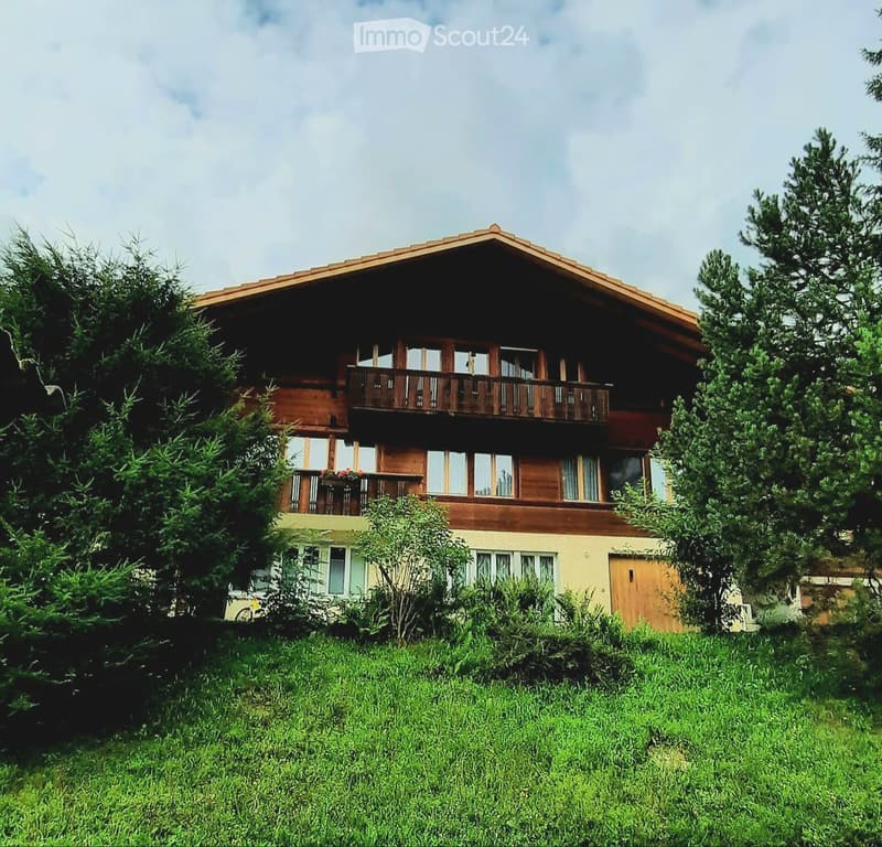 Two-story wooden house with balconies, surrounded by greenery and trees.