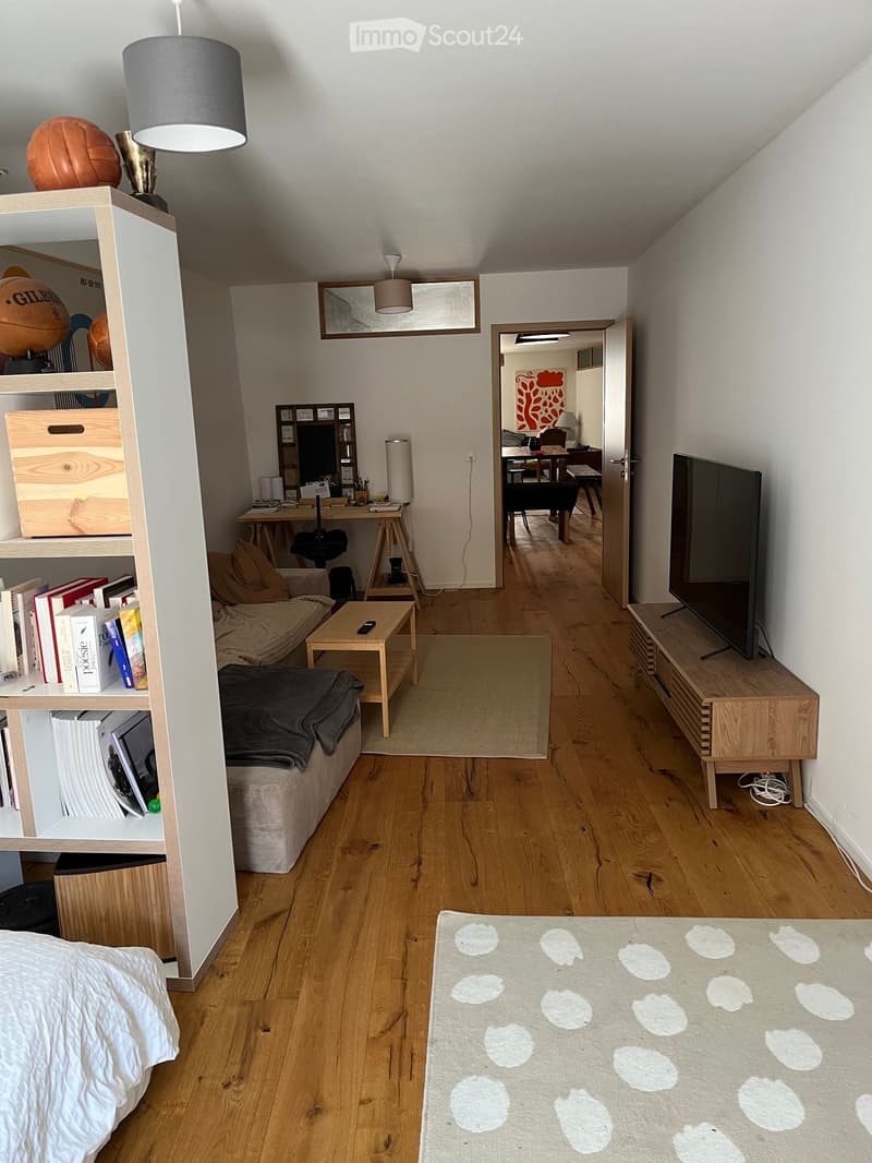 A modern living room with wooden flooring, a sofa, coffee table, bookshelf, and a TV. The room features a cozy seating area with a blanket and a small table, along with a workspace area visible through an open doorway.