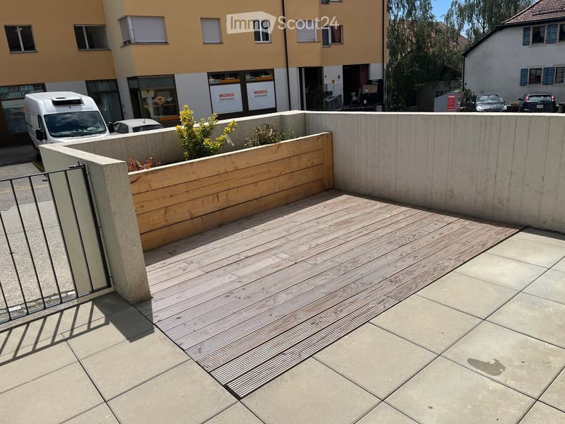 A modern outdoor patio with a wooden deck, concrete walls, and small plants. The area is enclosed by a metal gate and features a tiled floor.