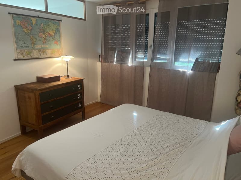 Cozy bedroom with a wooden bed, white bedding, a nightstand with a lamp, a wooden drawer unit, and a world map on the wall.