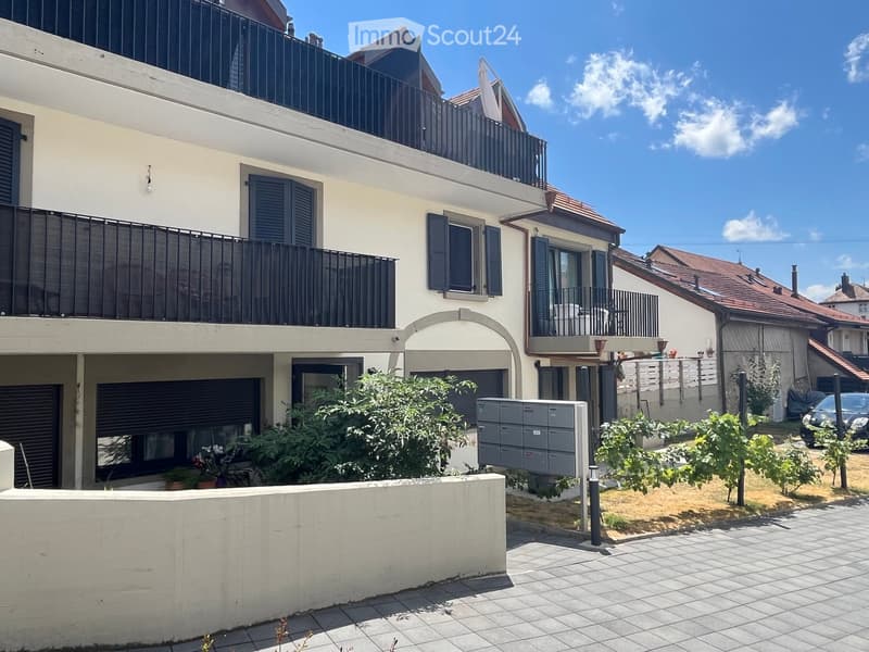 Two-story residential building with balconies, shutters, and a small garden area with potted plants and a mailbox.