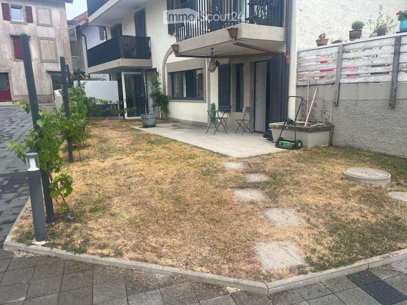 Garden area with dry grass, small tree, patio with chairs, and a building with balconies in the background.