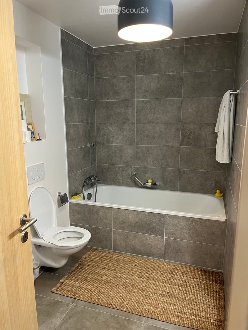 Modern bathroom with grey tiled walls, a white bathtub, a toilet, a towel hanging on the wall, and a woven bath mat on the floor.