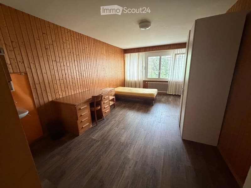 Bedroom with wooden walls and flooring, featuring a bed, desk, chair, and drawer unit. Large windows with blinds and curtains provide natural light.