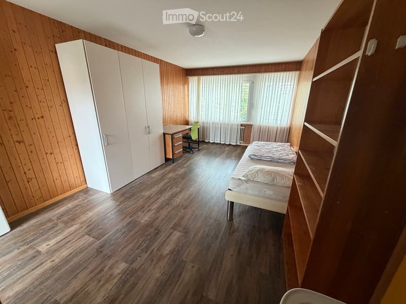 A furnished room with wooden walls and flooring, featuring a bed, desk, chair, and wardrobe.