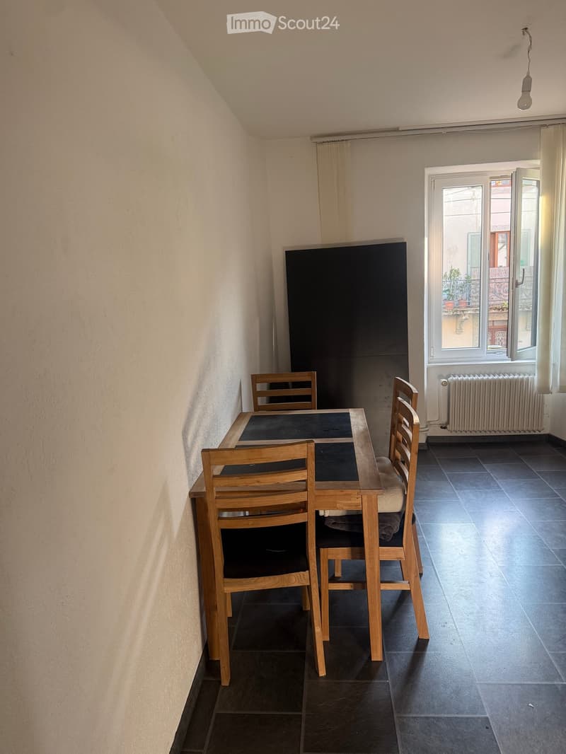 A small, furnished room with a wooden dining table and four matching chairs, dark tiled floor, and a large window with a view of a balcony.