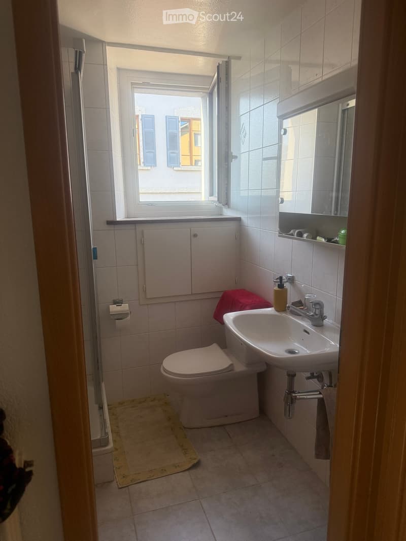 Small bathroom with white tiles, a toilet, a sink with a mirror above it, and a window with shutters.