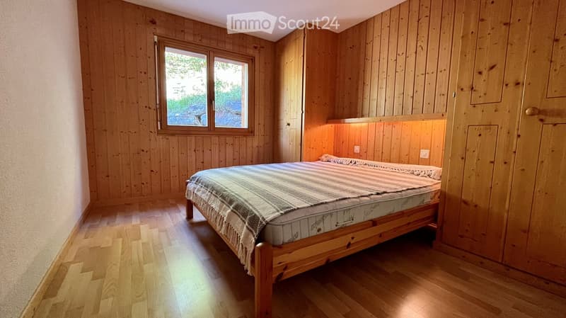 Cozy wooden bedroom with a single bed, wooden walls, and a large window offering a view of greenery.