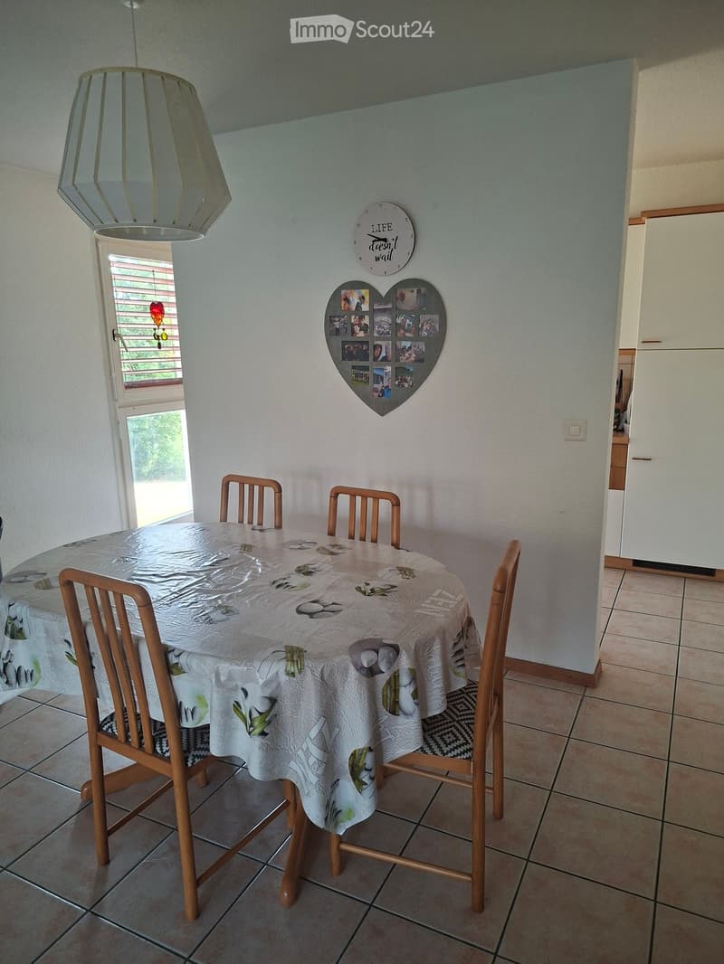 Dining area with a round table covered in a white tablecloth, surrounded by four wooden chairs. A heart-shaped collage of photos is mounted on the wall, and a modern clock with the inscription "Life doesn't wait" is visible. A refrigerator is partially vi