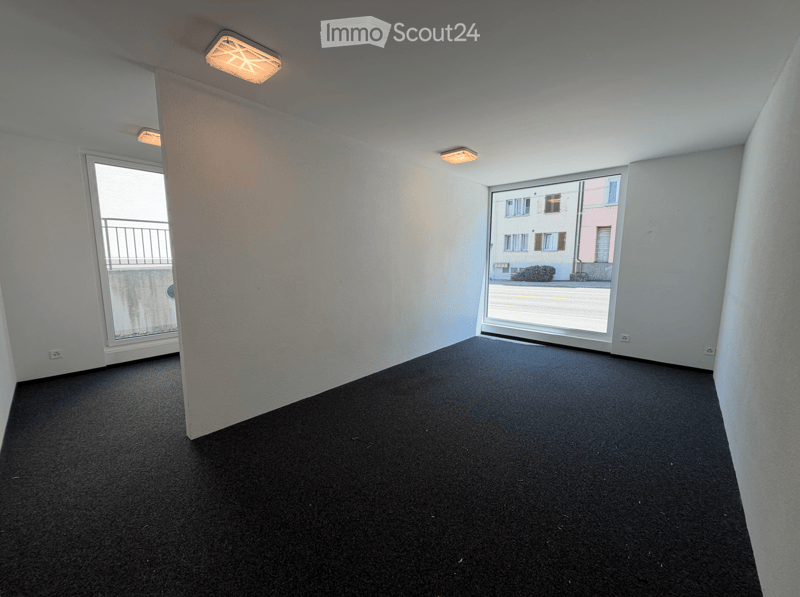 Empty room with white walls, black carpet, and large windows offering a view of the street and neighboring buildings.