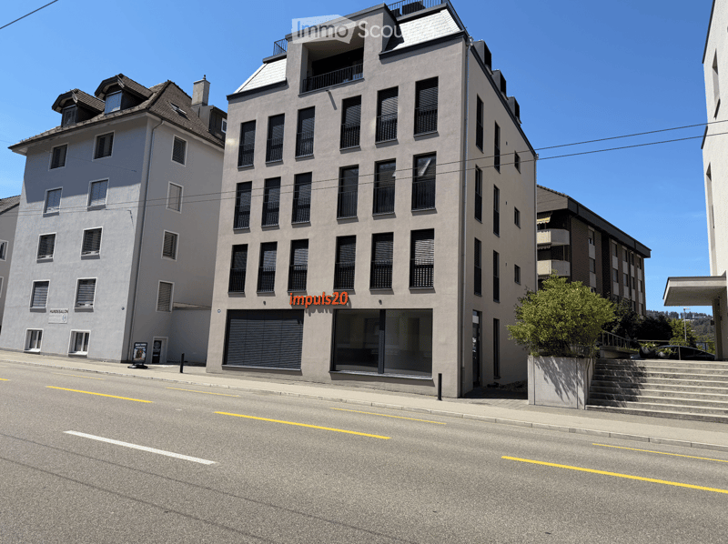 Modern multi-story building with large windows and balconies, located on a street with a clear blue sky and adjacent structures.