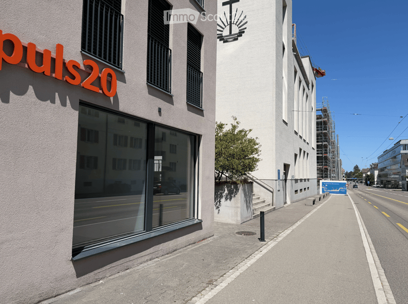 Modern building facade with large windows and a sign reading "puls20" in orange letters. The building has a clean, contemporary design with a small staircase leading up to the entrance.