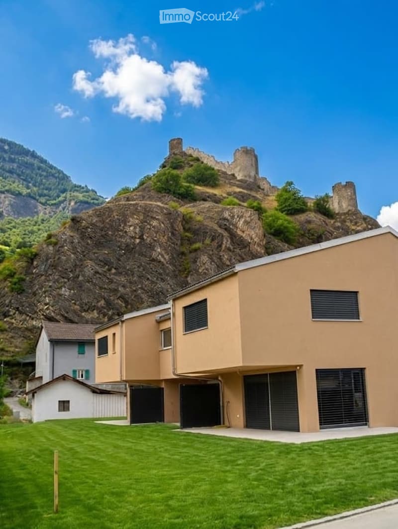 Modern two-story building with beige facade, large garage doors, and a well-maintained lawn, situated in front of a rocky hill with a castle-like structure on top.