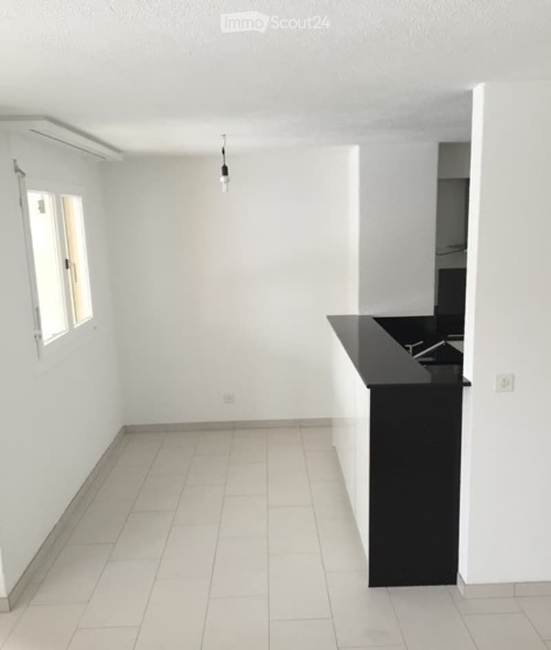 Empty room with white tiled flooring, white walls, and a small black countertop with a sink. There is a window on one wall and a light fixture hanging from the ceiling.