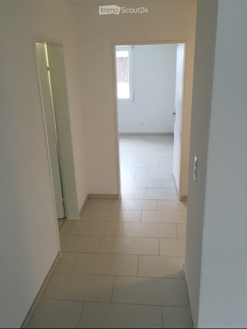 Empty hallway with tiled floor and white walls, leading to an open doorway with a window visible at the end.