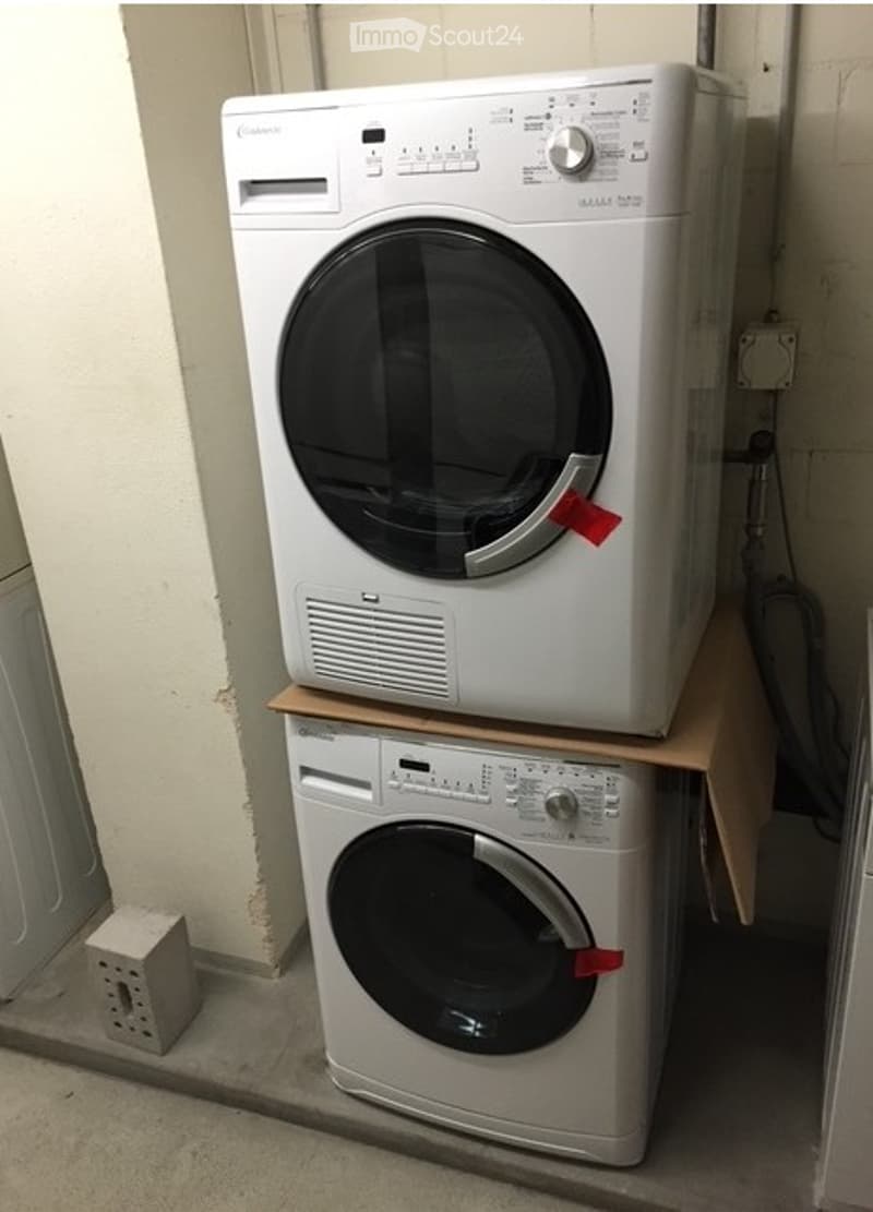 A small utility room with a washing machine and dryer stacked on a wooden platform, with a power outlet visible on the floor.
