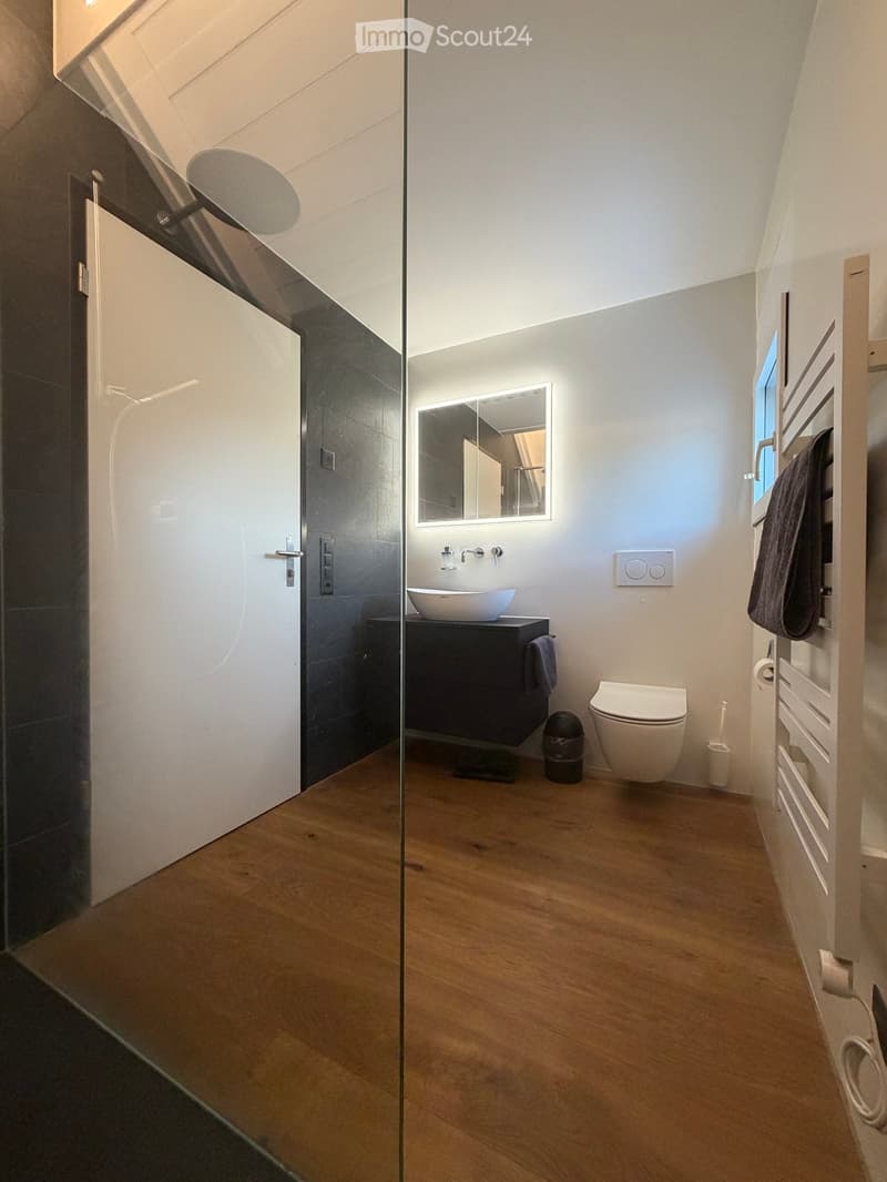 Modern bathroom with wooden flooring, a white sink on a dark cabinet, a toilet, a large mirror with LED lighting, and a towel on a heated towel rack.
