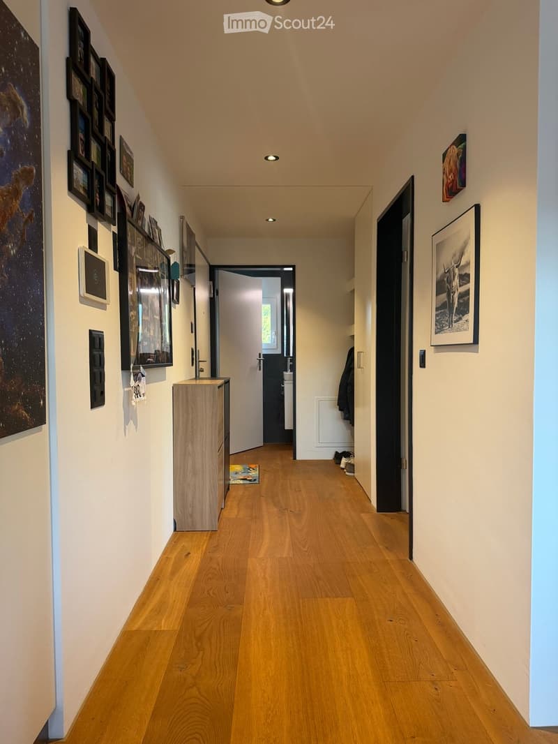 Modern hallway with wooden flooring, white walls, and several framed artworks. Features recessed lighting, a small wooden cabinet, and a partially open door leading to another room.