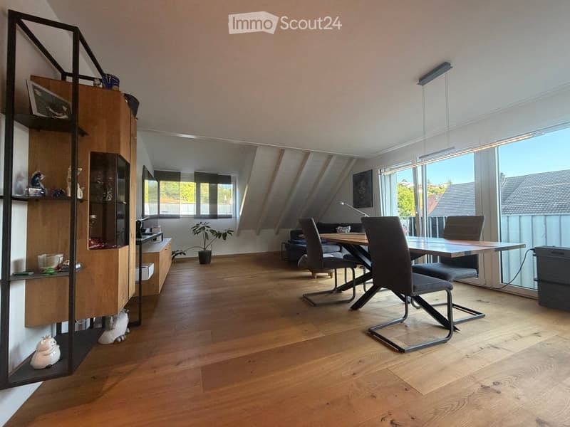 Bright, modern room with wooden flooring, large windows, and a mix of contemporary furniture including a dining table, chairs, and a shelving unit.
