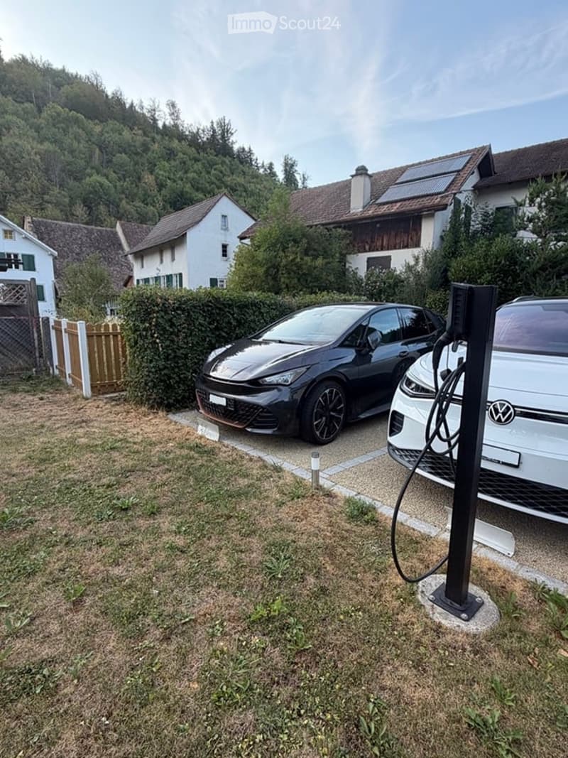 Residential area with two parked cars, one connected to a charging station, surrounded by houses and greenery.