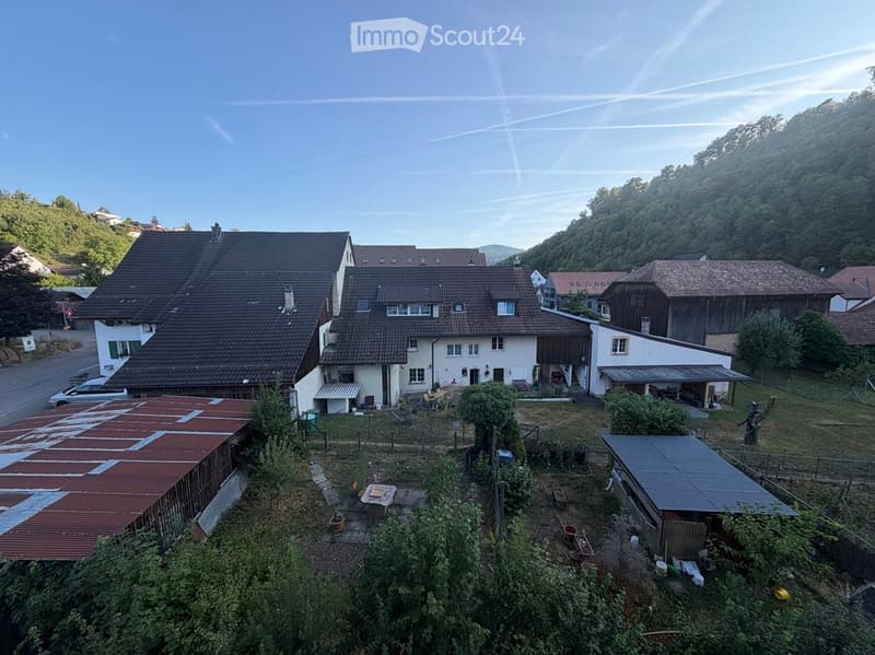 Aerial view of a residential area with multiple houses, surrounded by greenery and hills. The houses have pitched roofs and are set in a rural landscape.