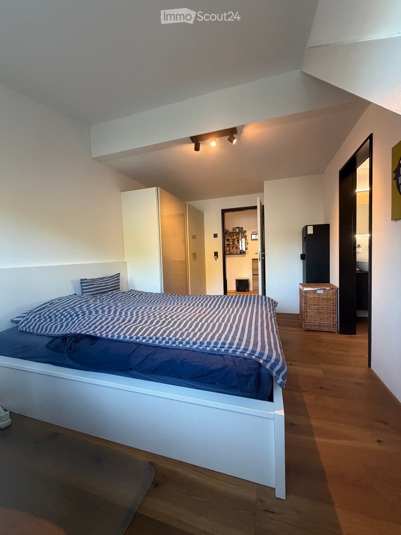 Modern bedroom with a white bed, striped bedding, wooden floor, wardrobe, and visible kitchen area with a refrigerator and wicker basket.