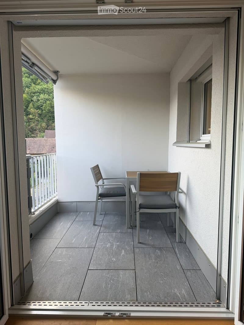 Balcony with grey tiled floor, white walls, two chairs and a small table, overlooking greenery.