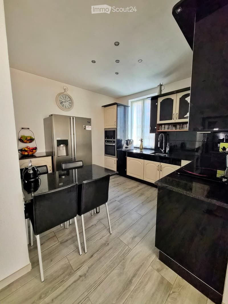 Modern kitchen with light-colored cabinets, dark countertops, and a dining area featuring a black table and chairs. Appliances include a refrigerator, oven, and blender, with a window and a fruit basket on the counter.