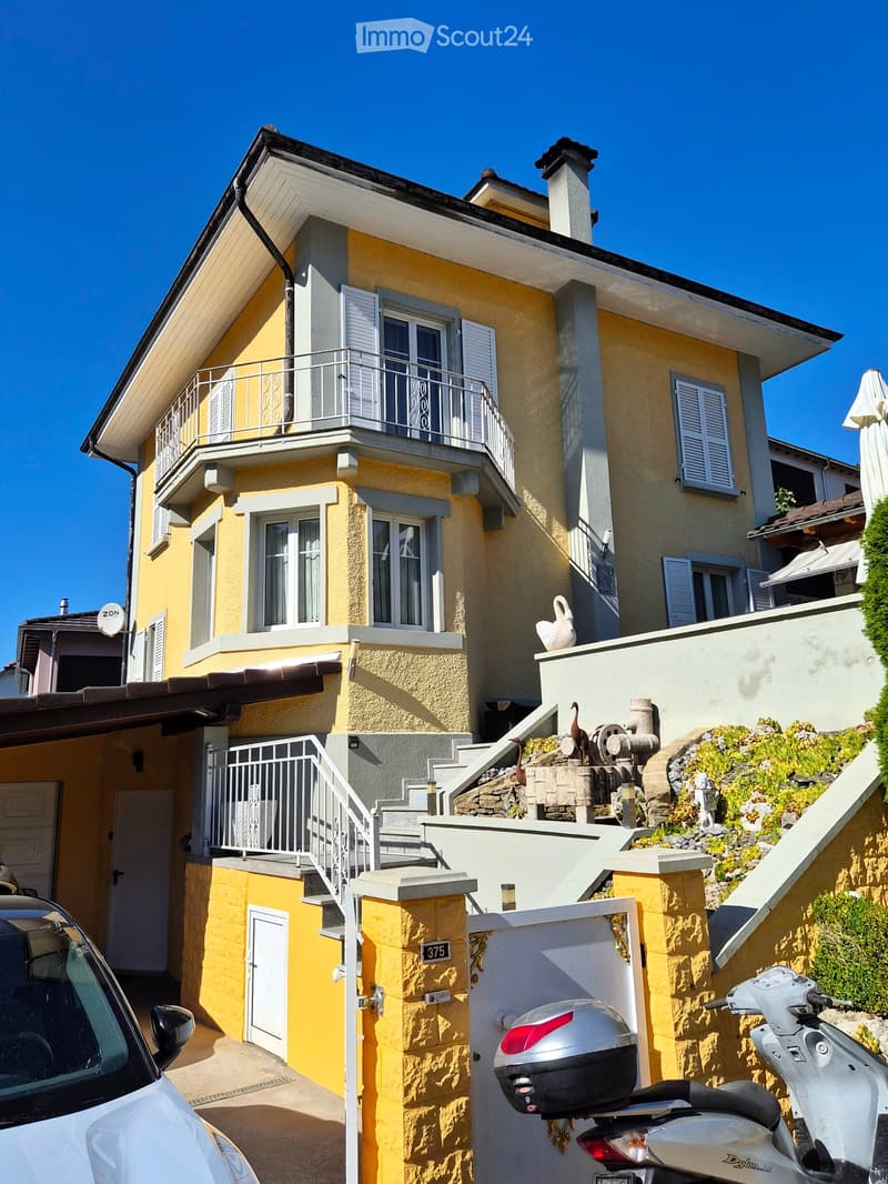 Two-story yellow house with white trim, multiple windows, and a balcony on the upper floor. A staircase leads to the entrance, and a scooter is parked nearby.