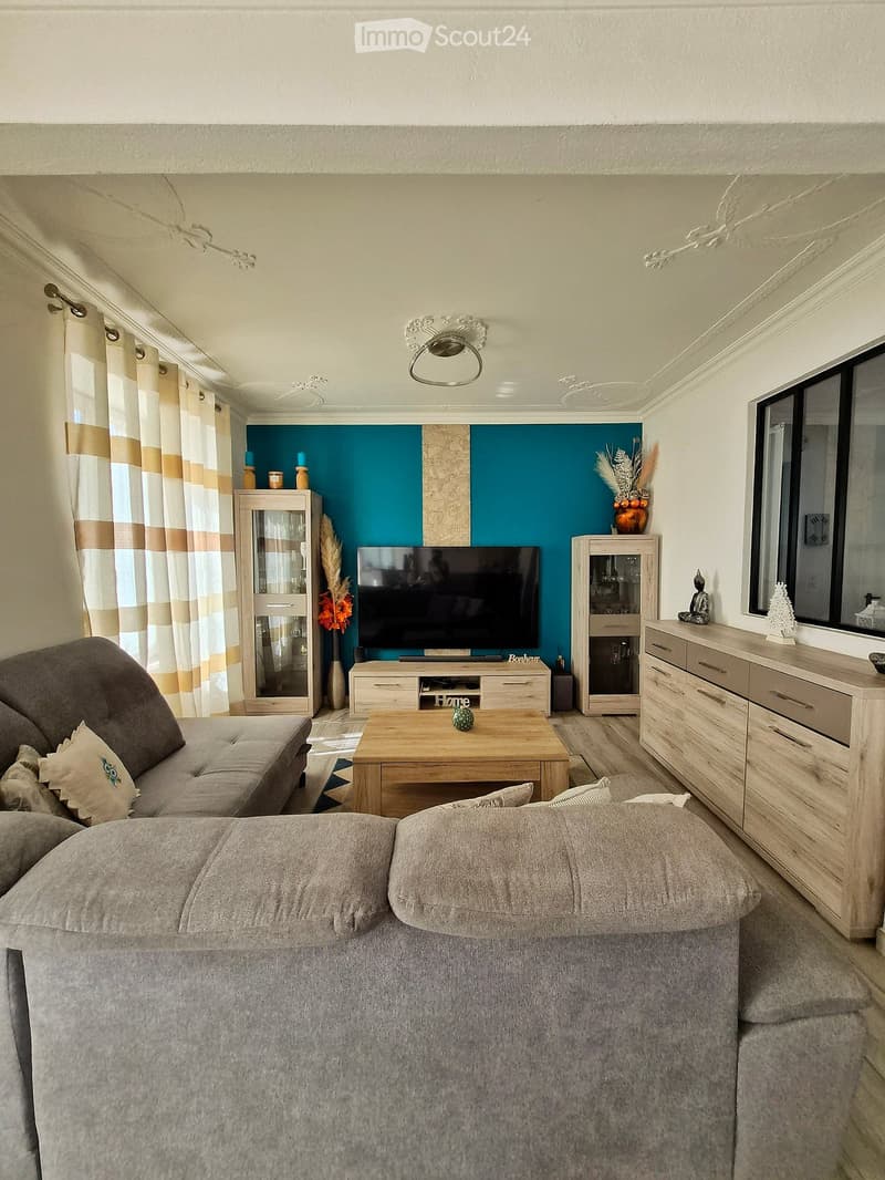 Modern living room with a grey sofa, wooden coffee table, flat-screen TV, and decorative cabinets. Features a blue accent wall, large window with curtains, and a vase with dried flowers on a side table.